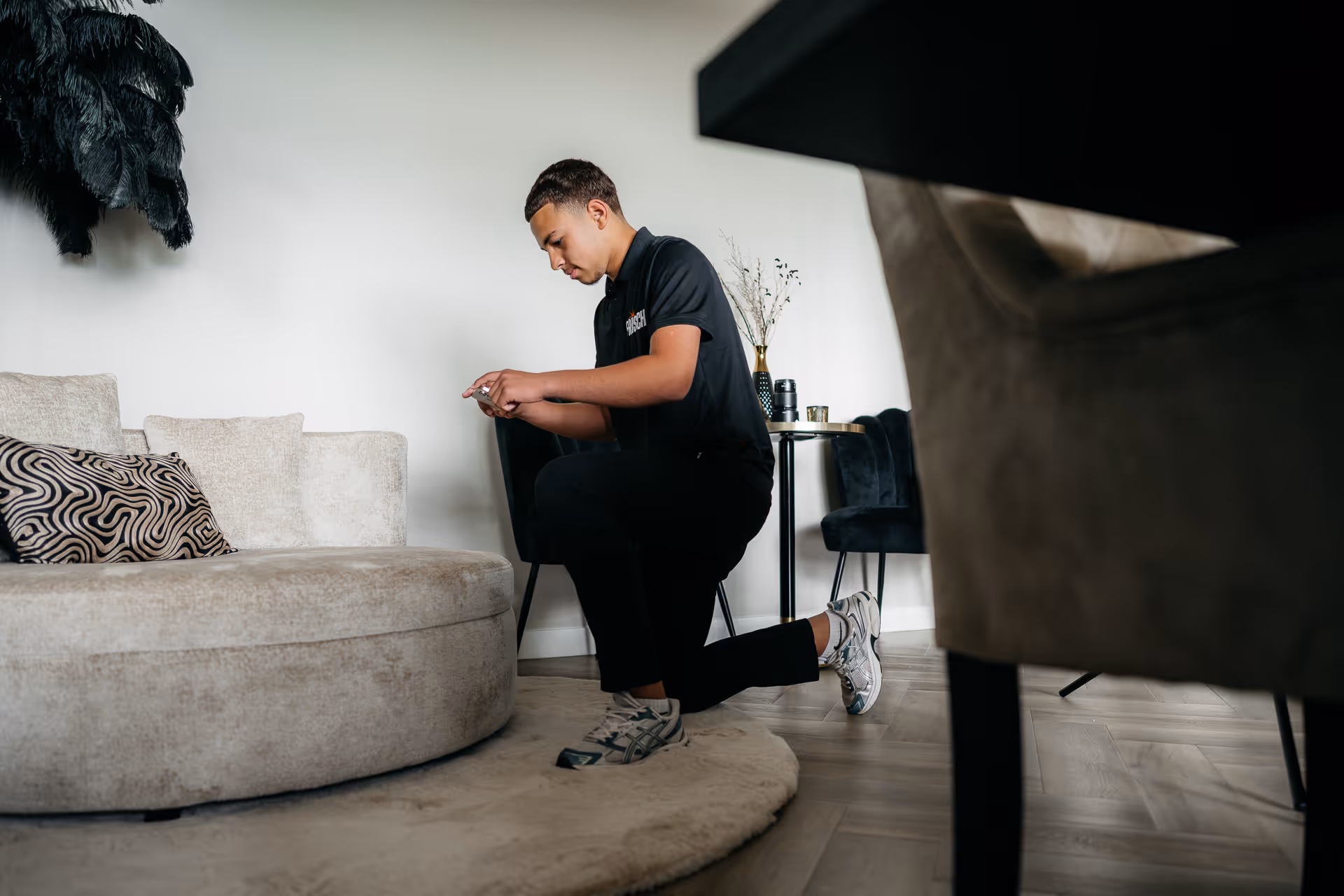 Jonge man knielt thuis op een ronde beige tapijt en gebruikt een smartphone.