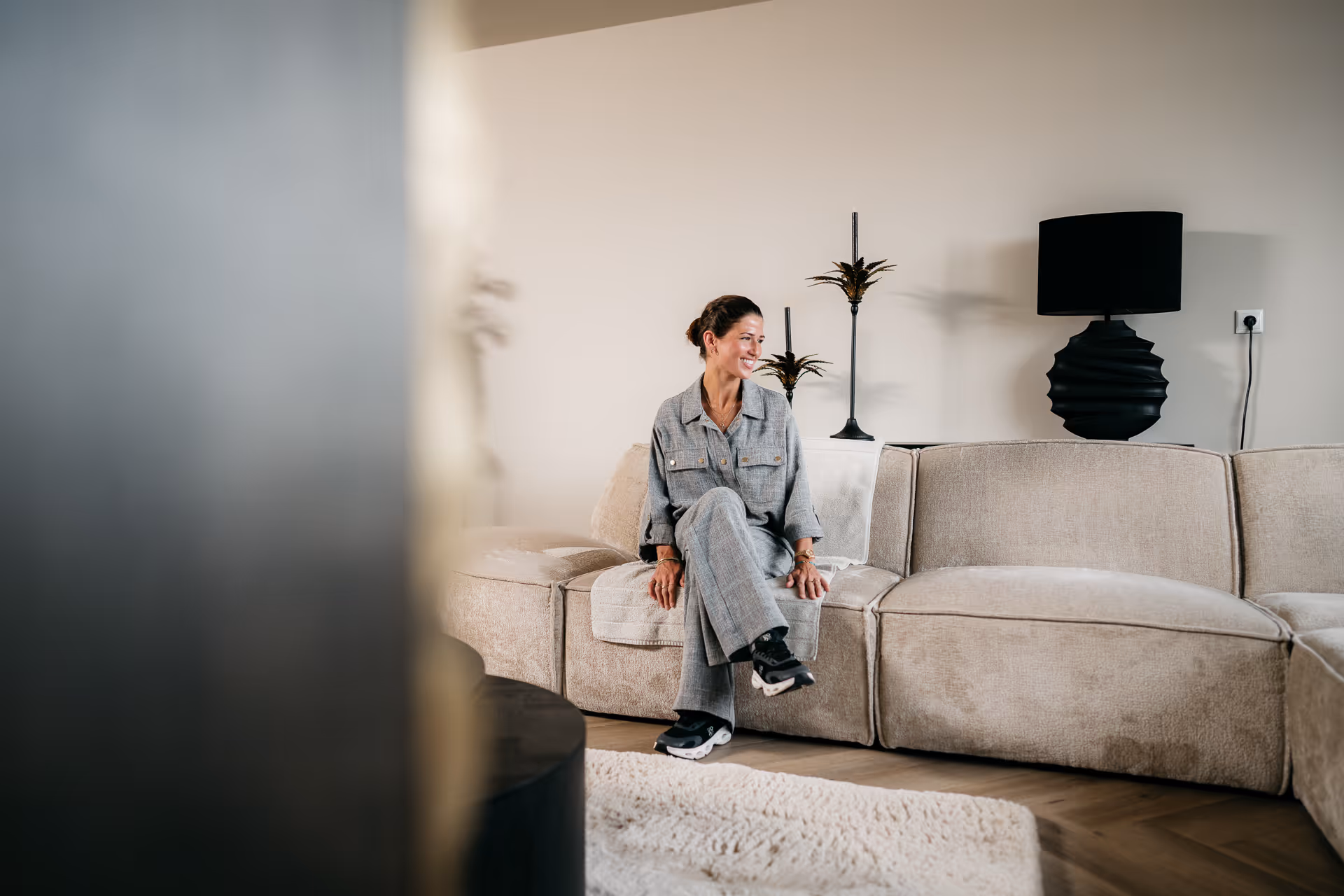 Vrouw in grijs pak zit op een beige bank in een moderne woonkamer met zwarte lampen.