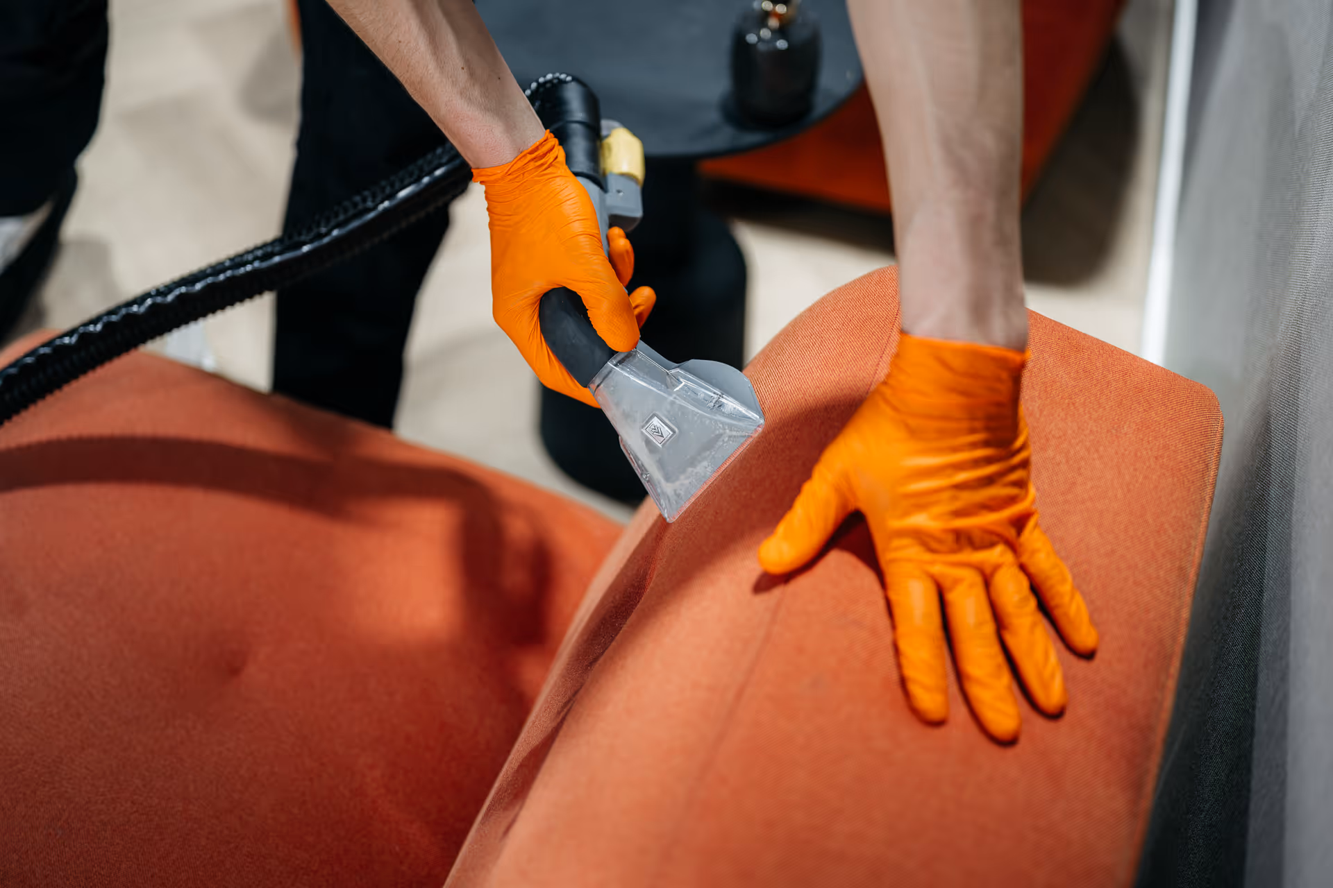 Hand met oranje handschoen reinigt een oranje stoffen stoel met een stoomreinigingsapparaat.
