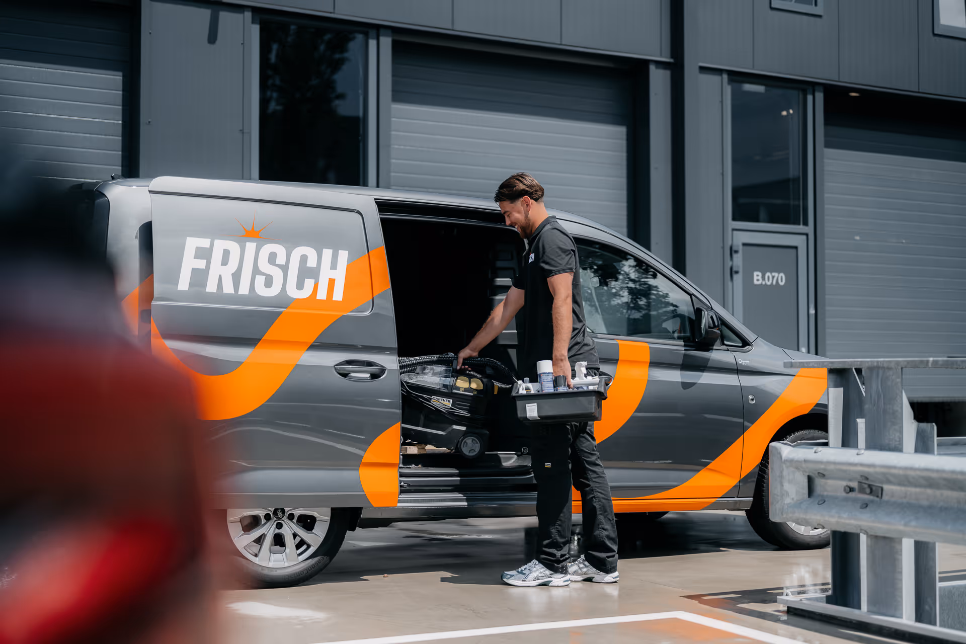 Man met mand met schoonmaakspullen bij een grijze bus met oranje accenten en tekst Frisch.
