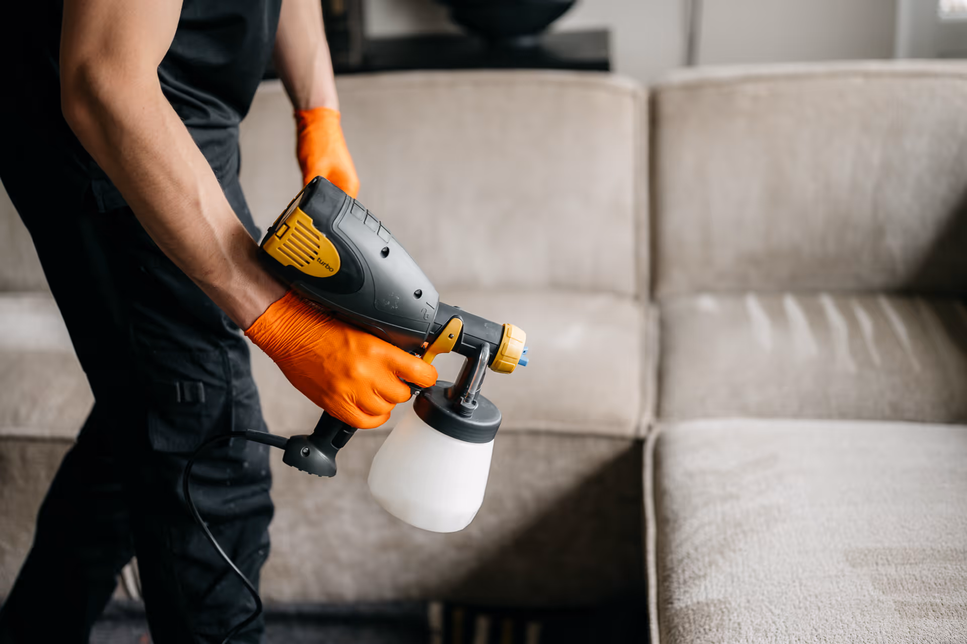 Persoon met oranje handschoenen gebruikt een elektrische spray om een beige bank te reinigen.