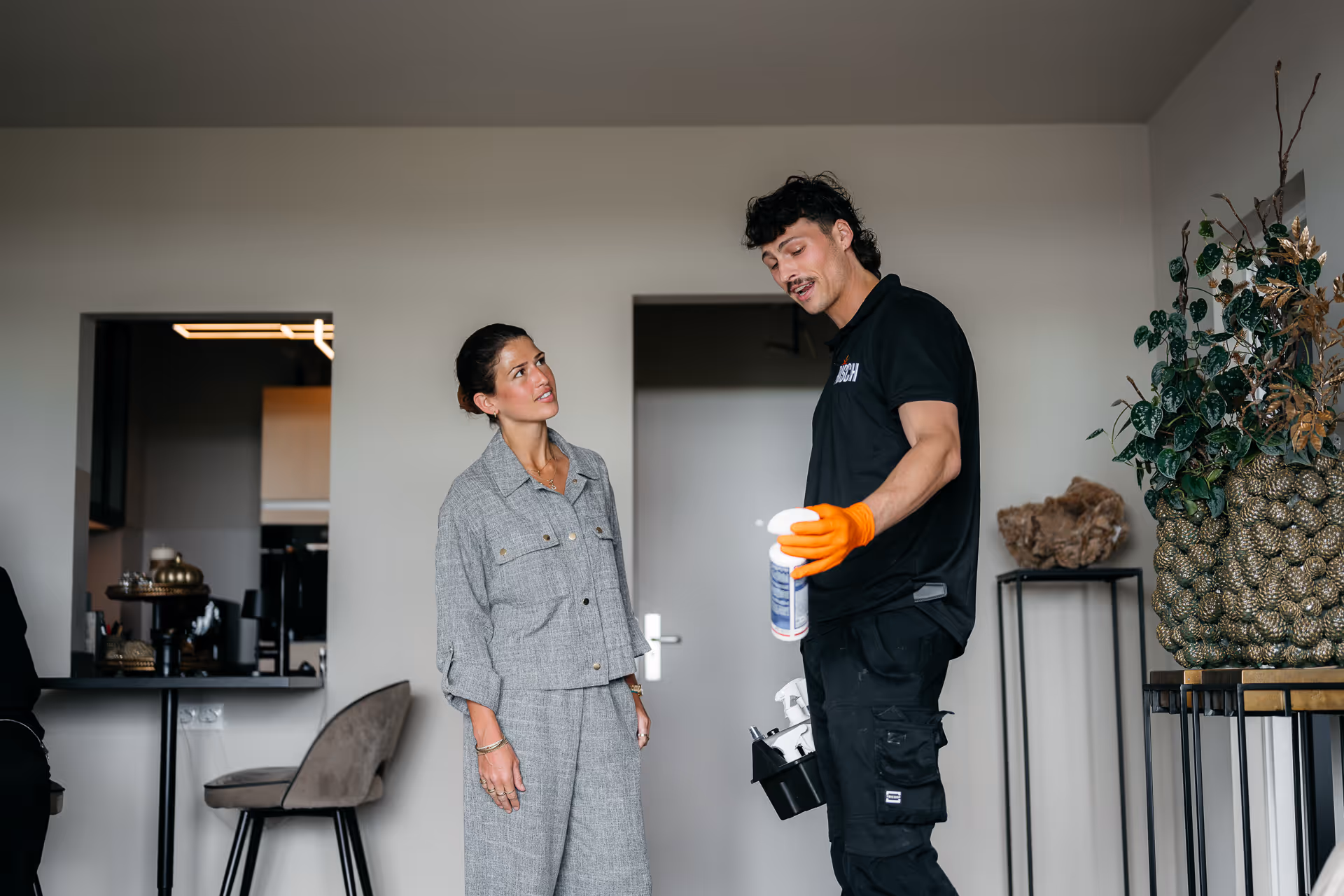 Man met handschoenen toont een schoonmaakproduct aan vrouw in een moderne kamer.