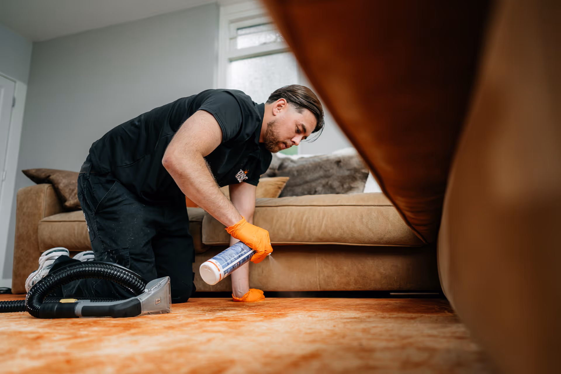 Man in oranje handschoenen reinigt tapijt naast een bruine bank in een lichte woonkamer.