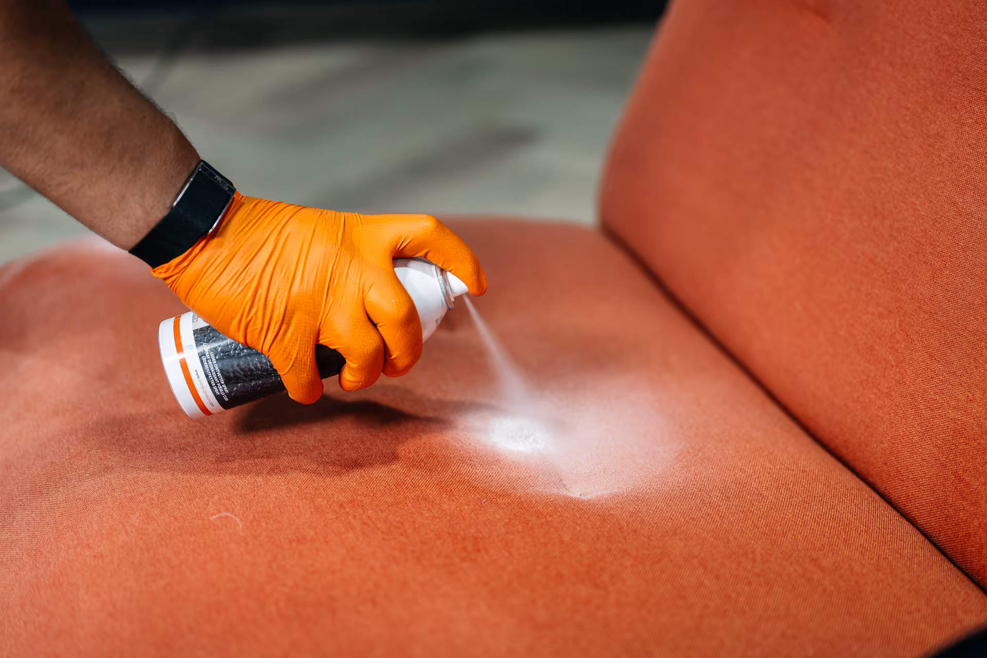 Hand with orange glove spraying foam cleaner on an orange fabric sofa seat.