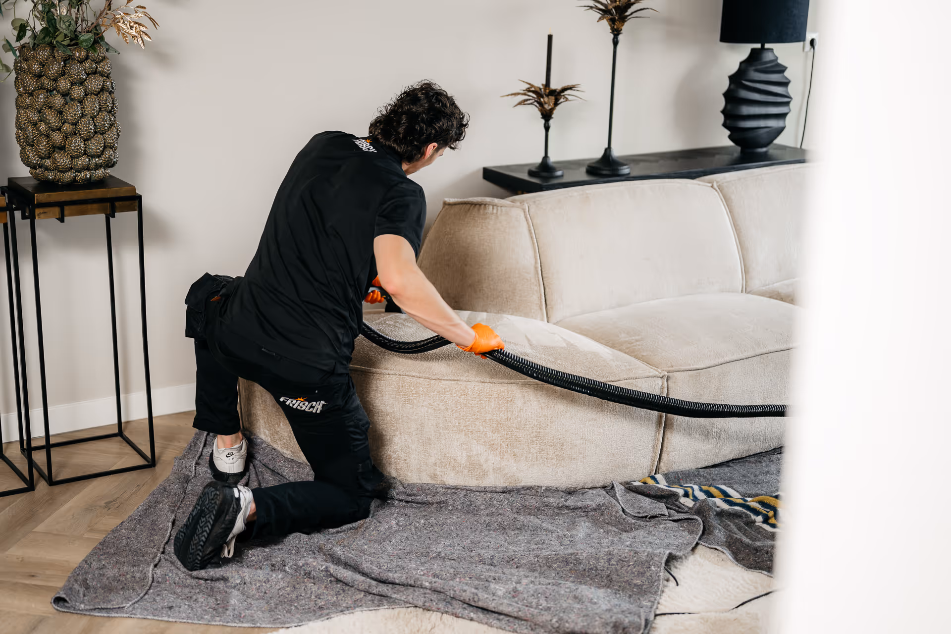 Man met oranje handschoenen reinigt een beige bank met een stofzuiger in een moderne woonkamer.
