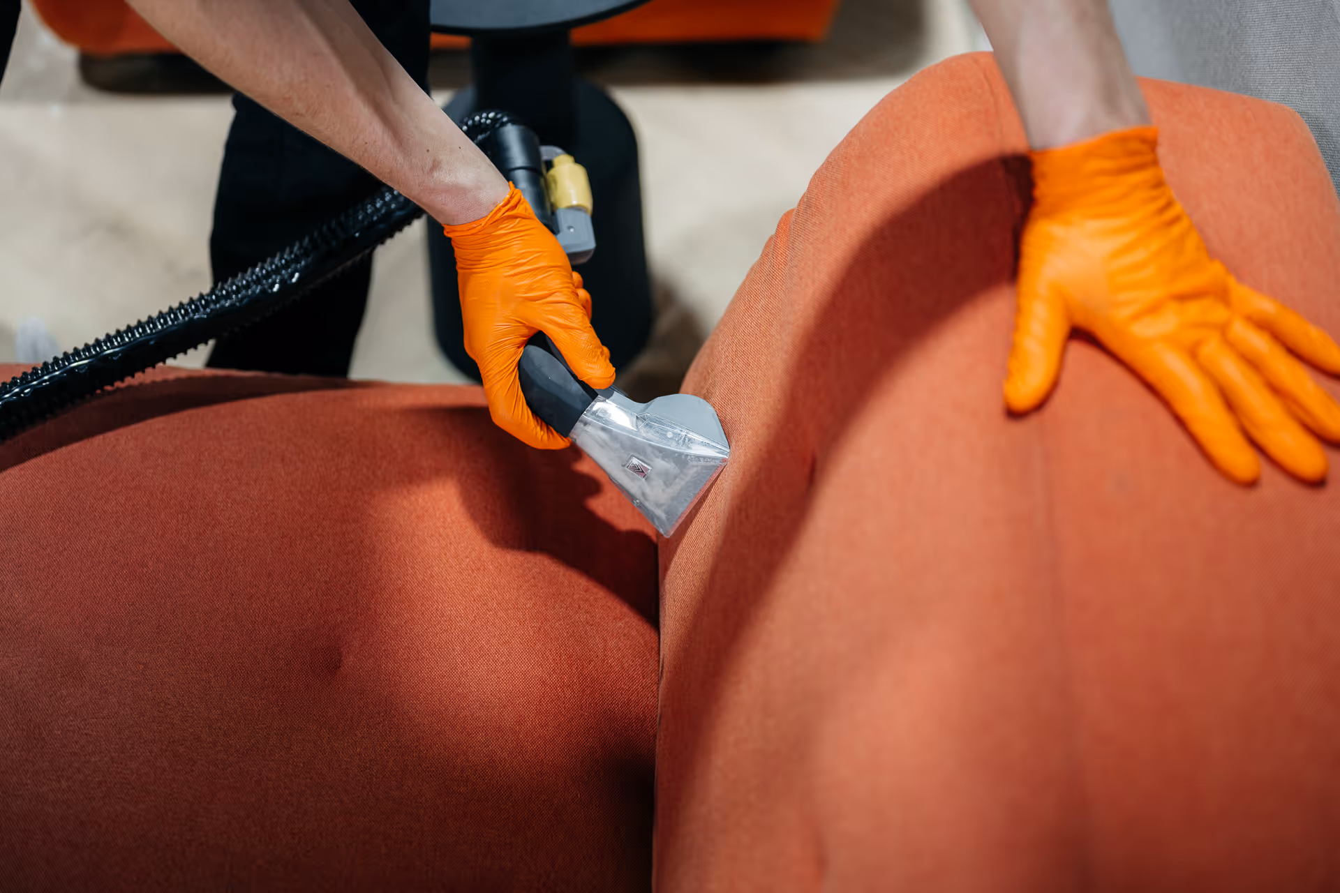 Persoon met oranje handschoenen reinigt oranje bank met stoomreinigingsapparaat.