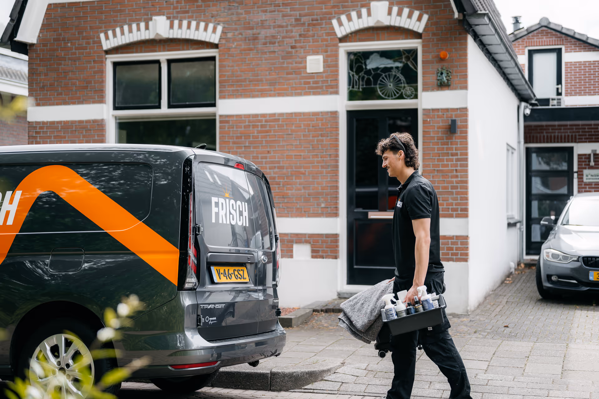 Man draagt schoonmaakartikelen en een deken lopend naar een bestelwagen met tekst 'FRISCH' in woonwijk.