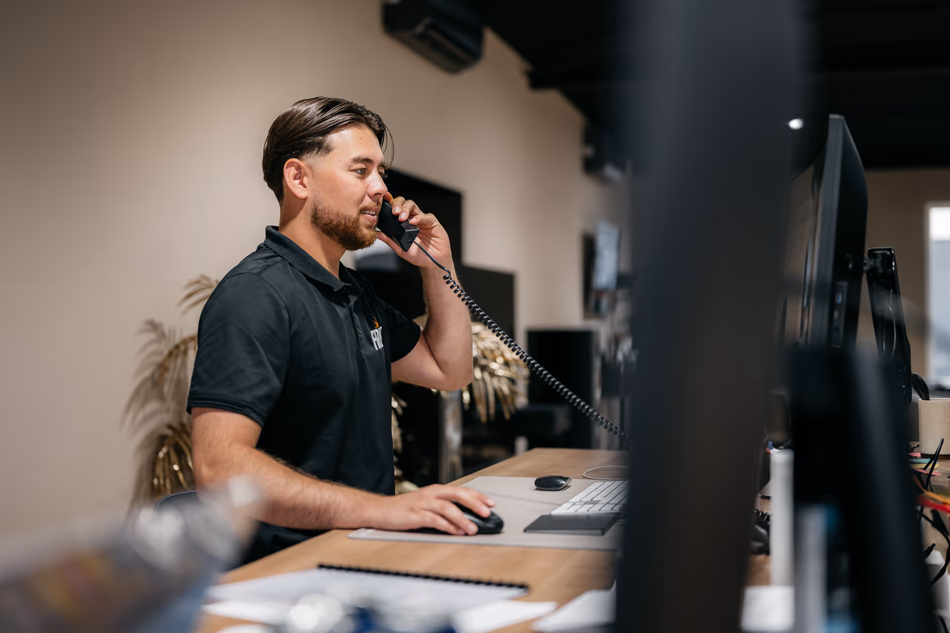 Man in black polo shirt talks on phone while working at desk with computer in modern office.