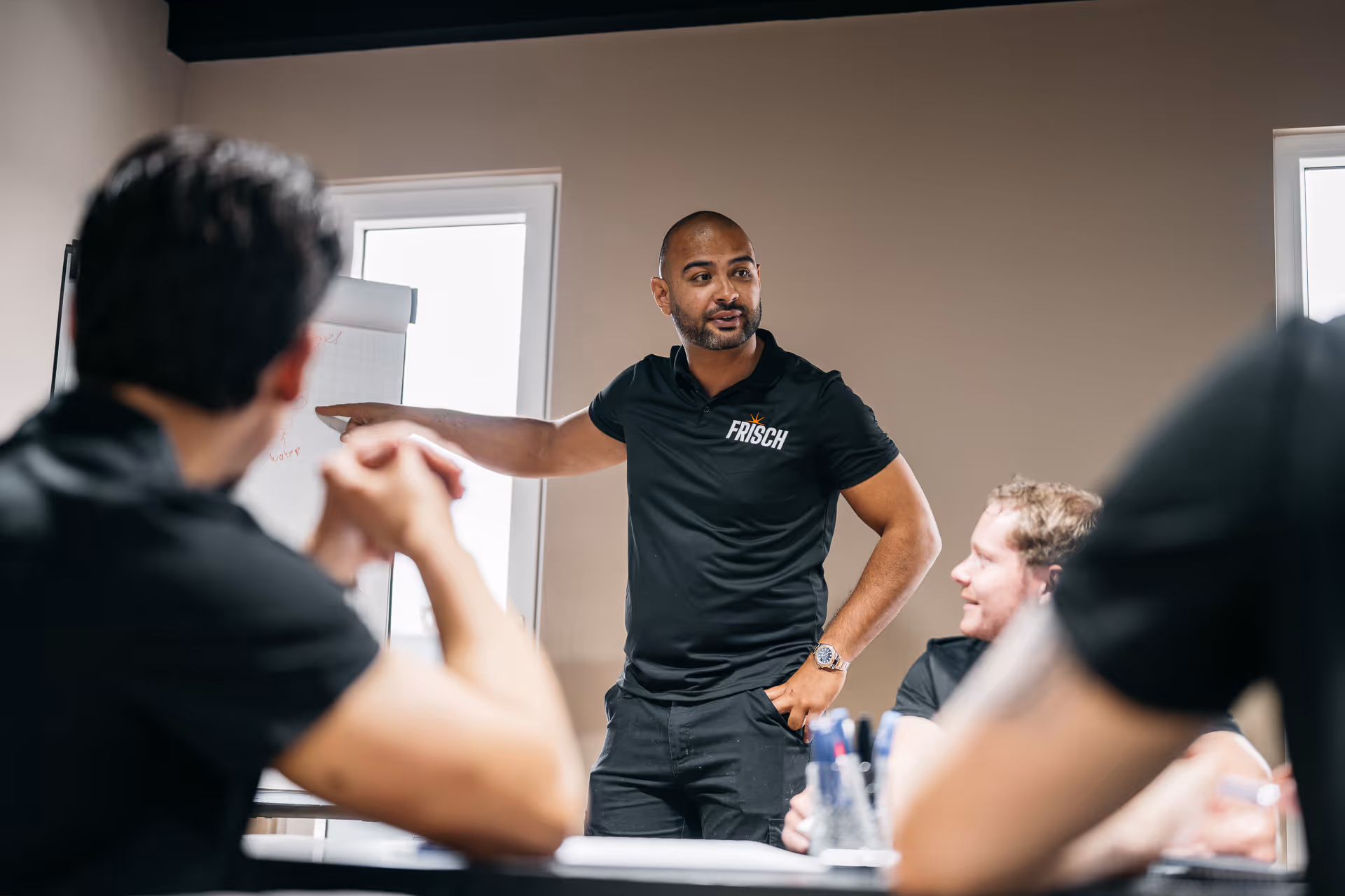 Man in black FRISCH shirt points to a flip chart during a meeting with seated colleagues.