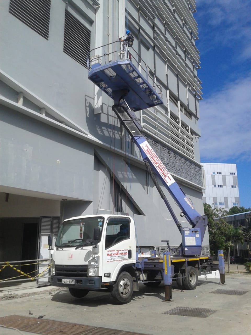 White truck with extended boom lift raising a worker in a blue safety helmet near a gray building.