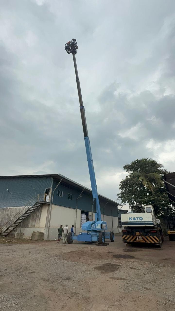 Blue aerial lift extended high near warehouse with three people and a KATO truck nearby.
