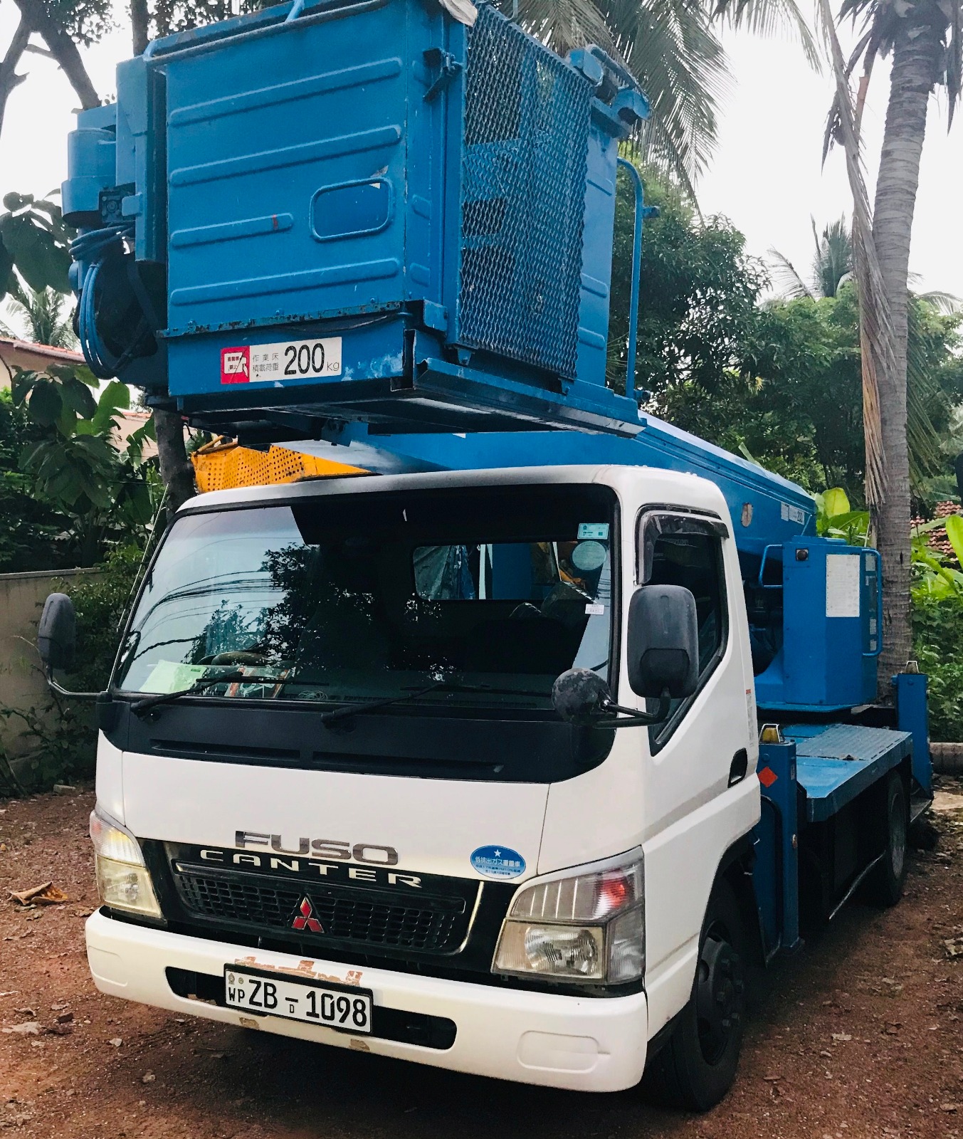 White Mitsubishi Fuso Canter truck with blue hydraulic lift equipment parked on dirt near palm trees.