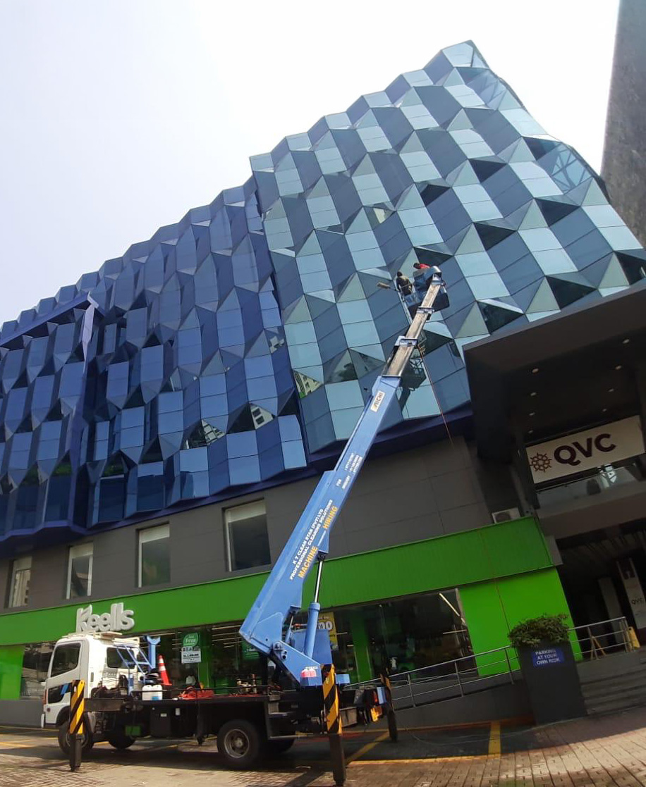Workers on a blue lift cleaning glass panels of a modern building with geometric façade above a Keells store.