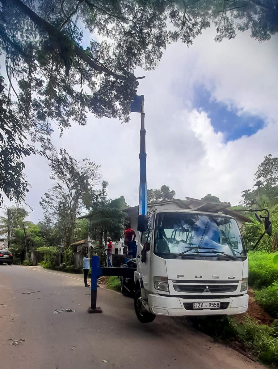 White Fuso truck with an extended blue crane pruning tree branches on a roadside.