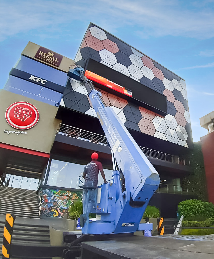 Worker on blue lift cleaning geometric patterned cladding on a commercial building with stores and theaters.