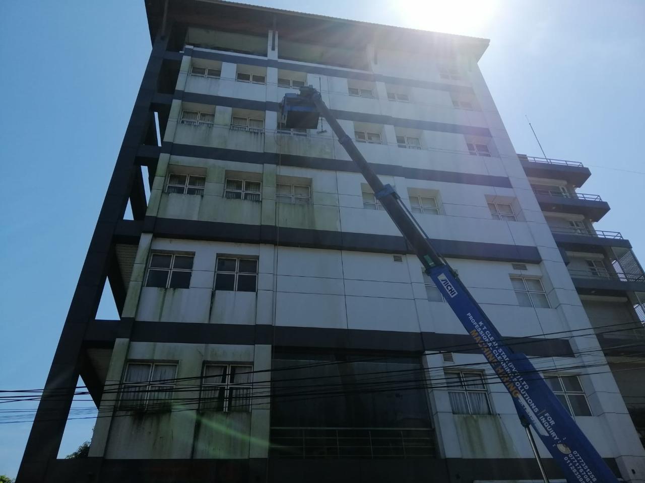 Worker on a blue hydraulic lift cleaning the stained exterior of a tall white building under bright sunlight.
