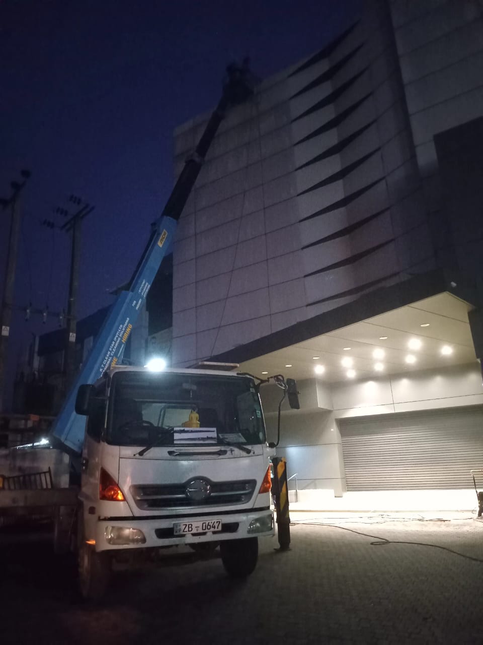 White truck with blue crane lifts worker cleaning cladding on a lit building at night.