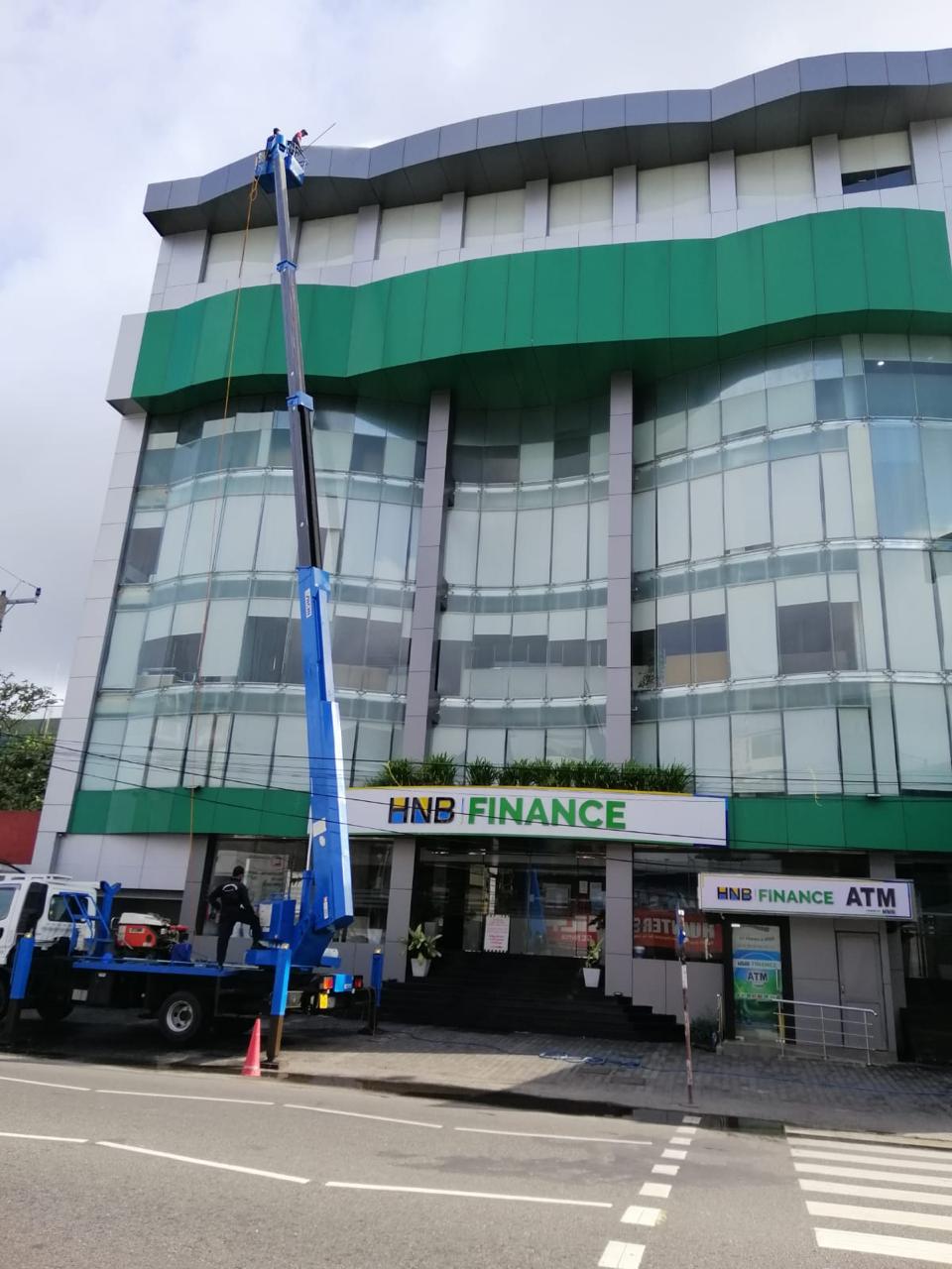 Worker in lift cleaning cladding on top of HNB Finance building with green and glass facade.