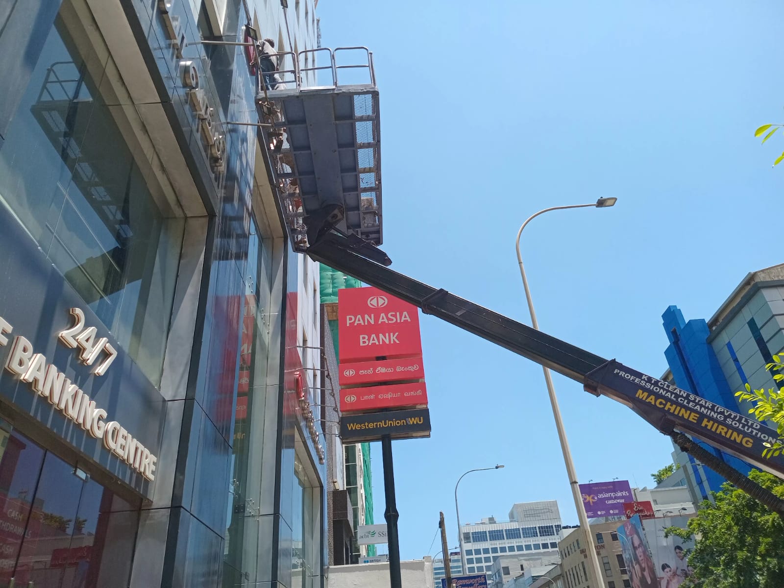 Worker on elevated platform cleaning glass windows of a banking center building under clear blue sky.