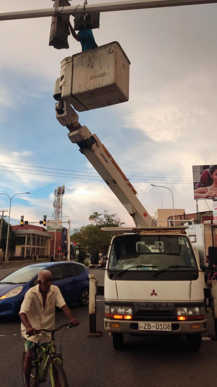 Worker in lifted bucket truck repairing a traffic light above a street with vehicles and cyclist below.