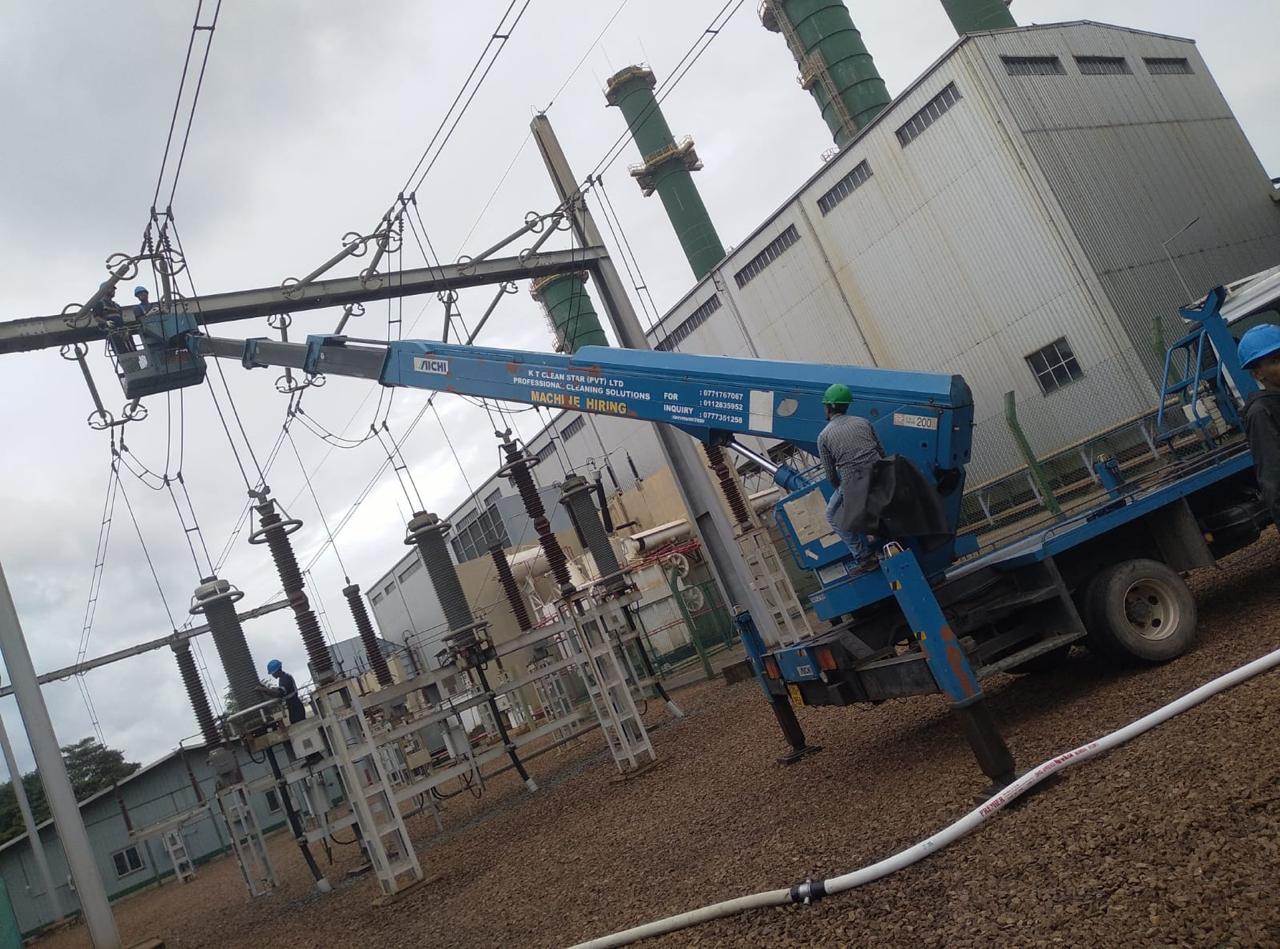 Workers in helmets operate a blue lift truck near electrical equipment and industrial buildings.