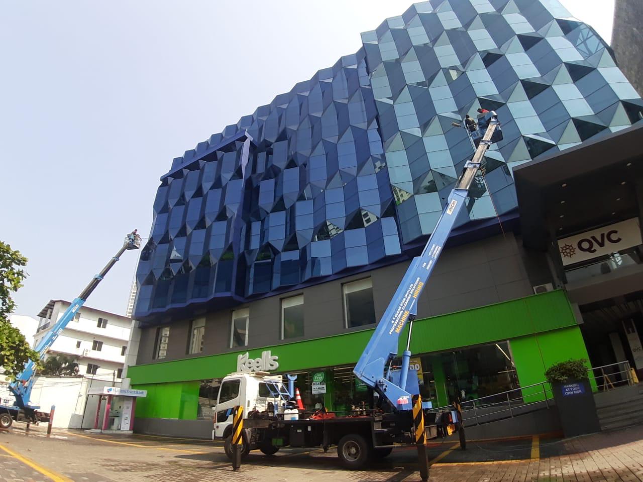 Two workers on lifts cleaning glass panels of a modern blue-tinted building facade.