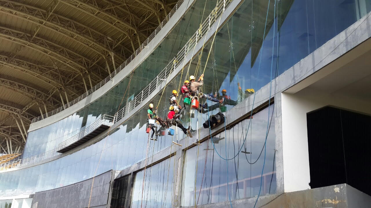 Workers wearing helmets suspended by ropes cleaning large curved glass windows on a building exterior.