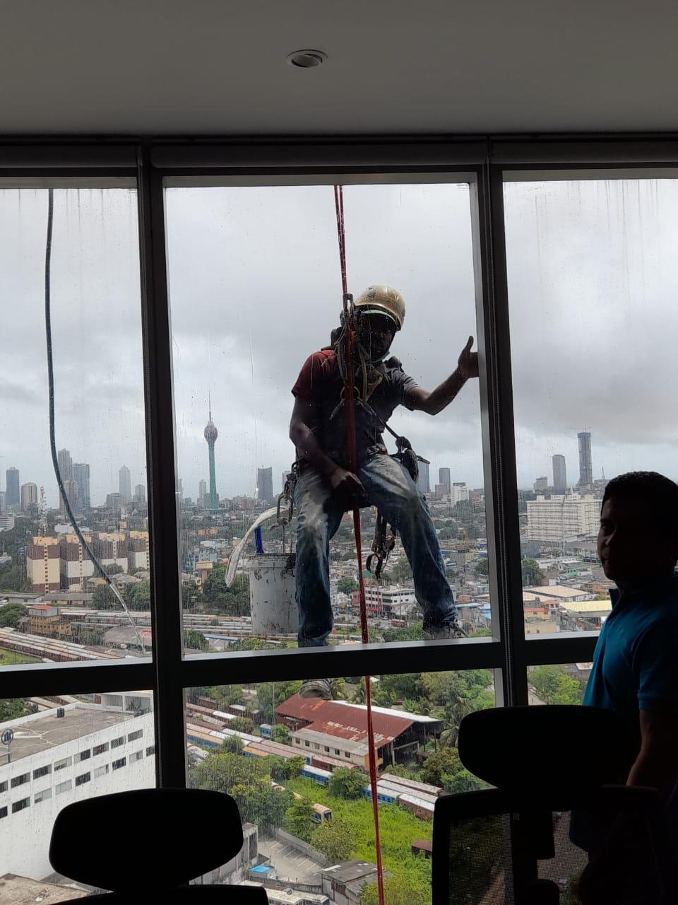 Window cleaner in safety gear suspended outside a high-rise building giving a thumbs up.