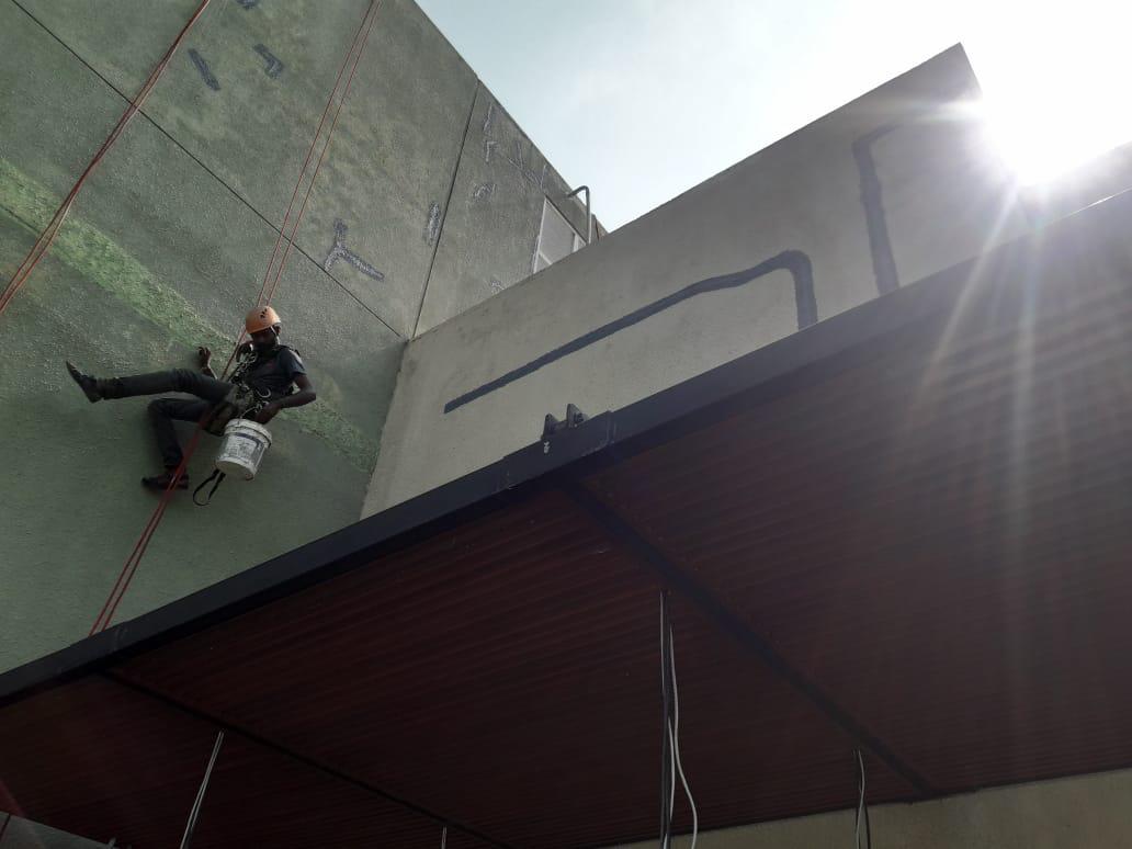 Worker wearing a harness and helmet rappelling down a building wall with a bucket, under bright sunlight.