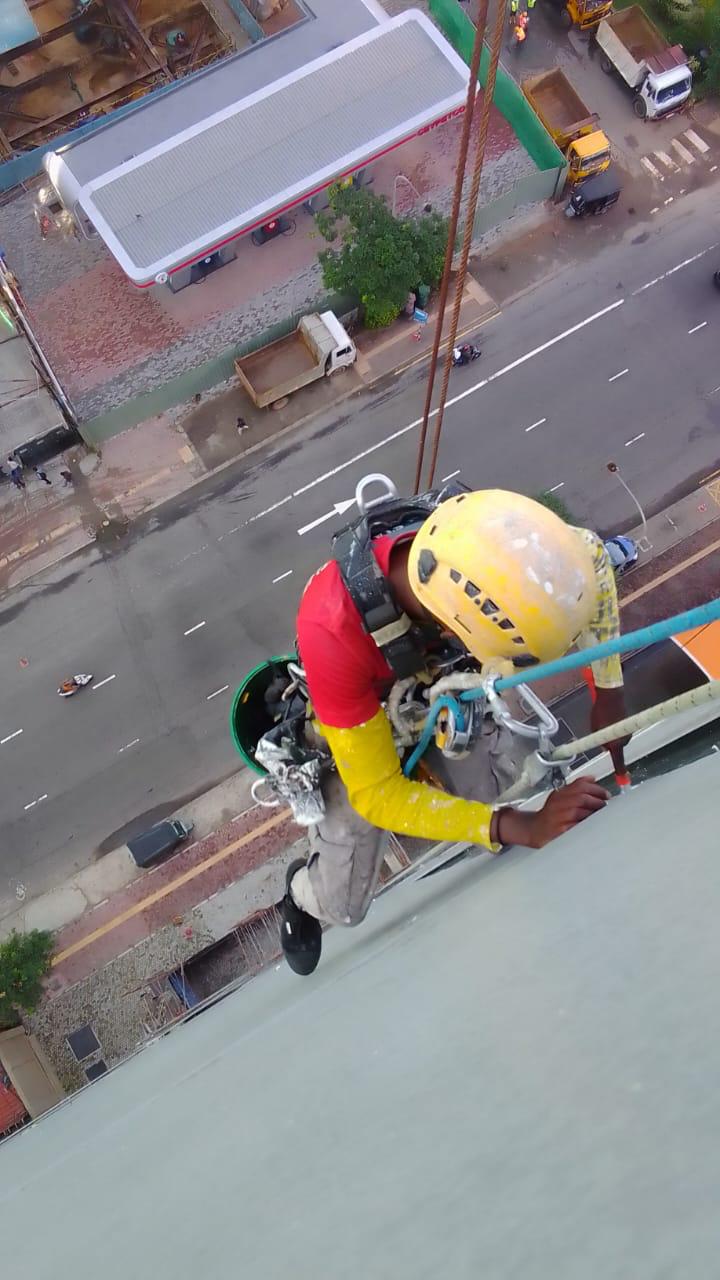 Worker in yellow helmet and red shirt rappelling down a tall building with climbing gear.