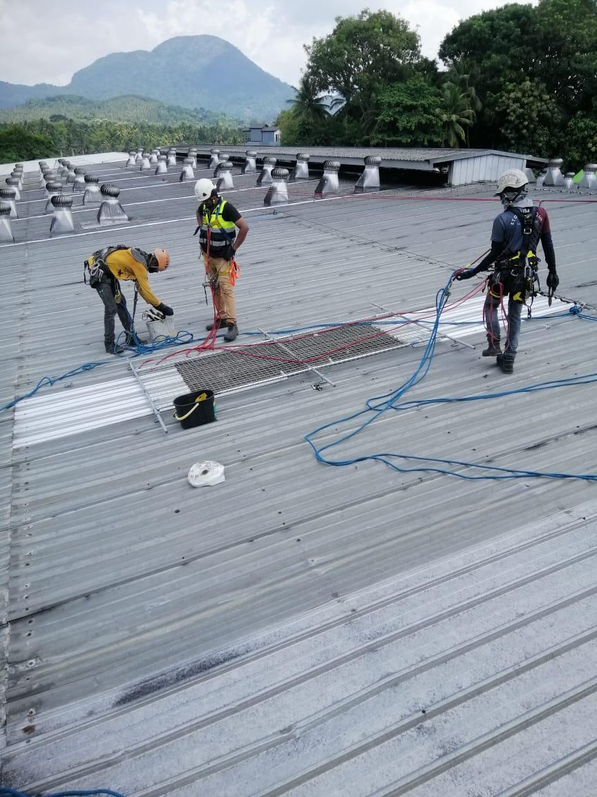 Three workers in safety gear securing ropes on a metal roof with vent pipes and a mountain in the background.