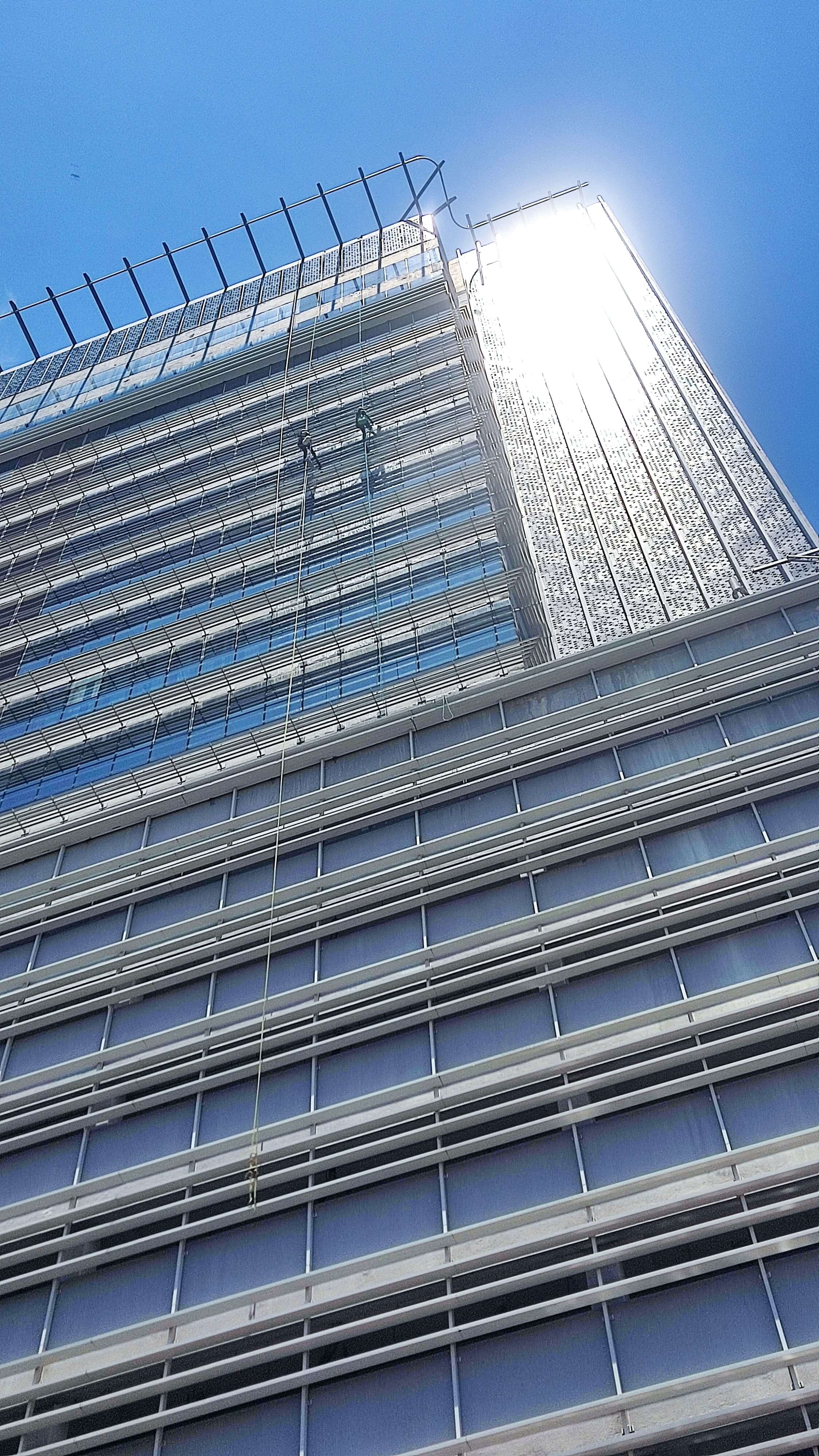 Two workers in safety gear perform rope access cleaning on a tall glass and metal building in sunlight.