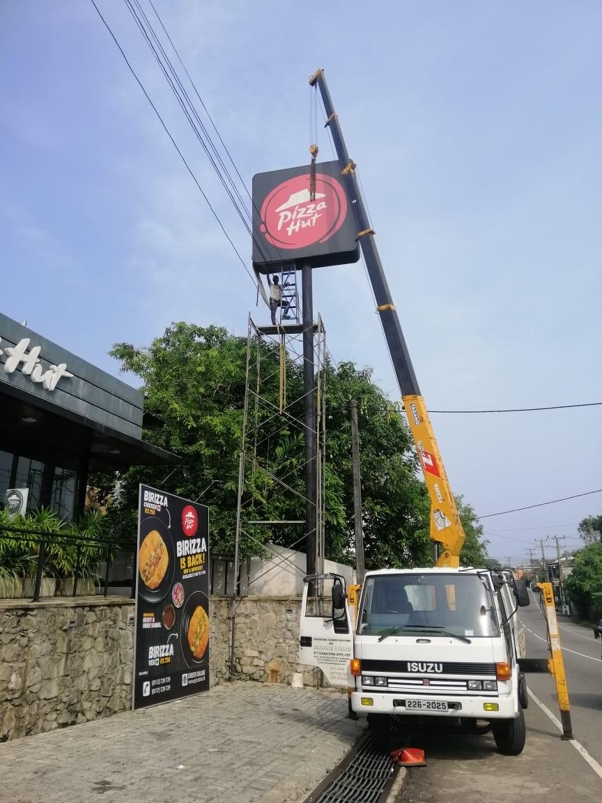 Crane truck with open door lifts worker to a tall Pizza Hut sign near a stone wall and food billboard.