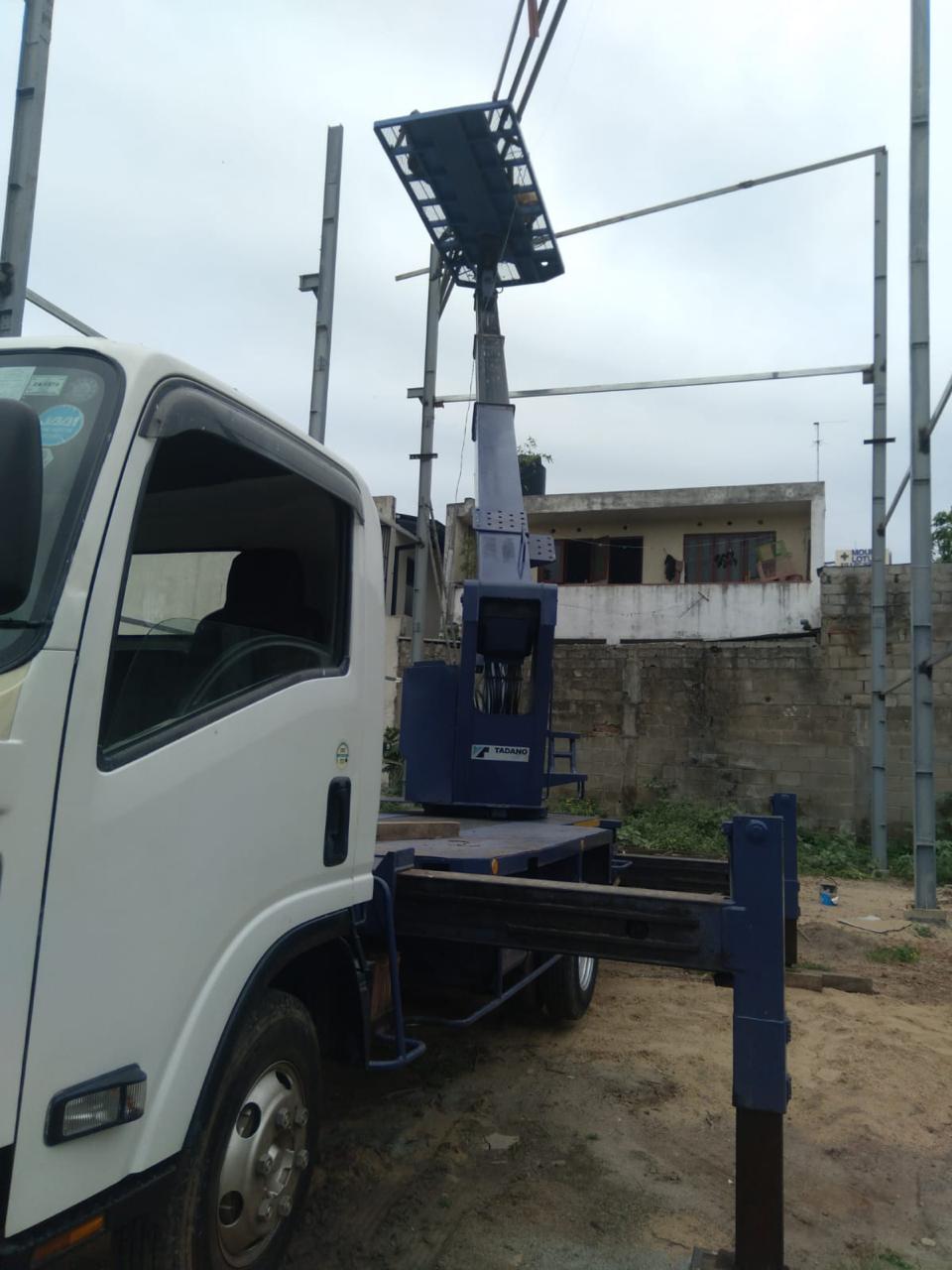 White truck equipped with a blue hydraulic crane parked on sandy ground near a concrete wall.
