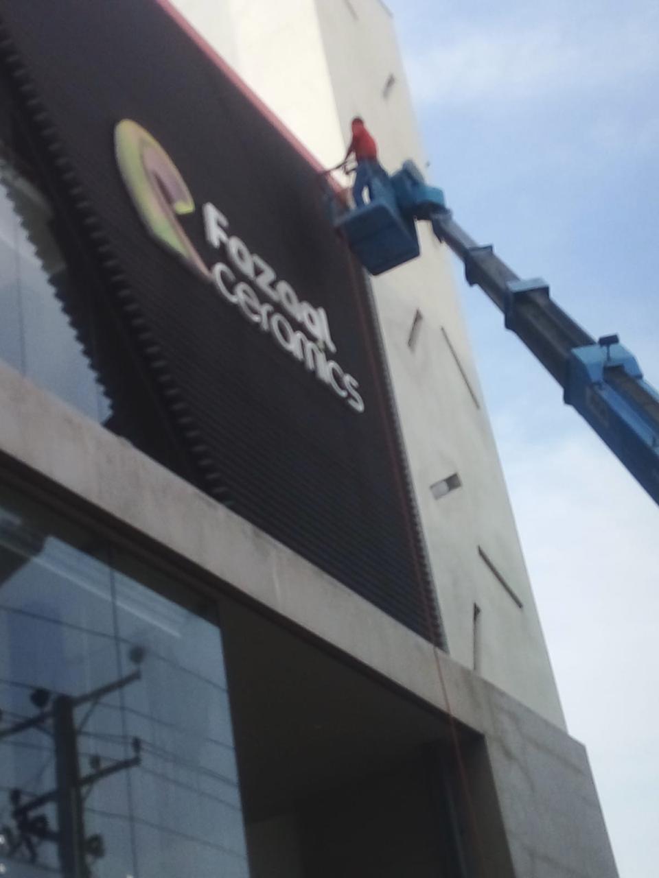 Worker on a blue cherry picker cleaning a large Fozzool Ceramics sign on a building.