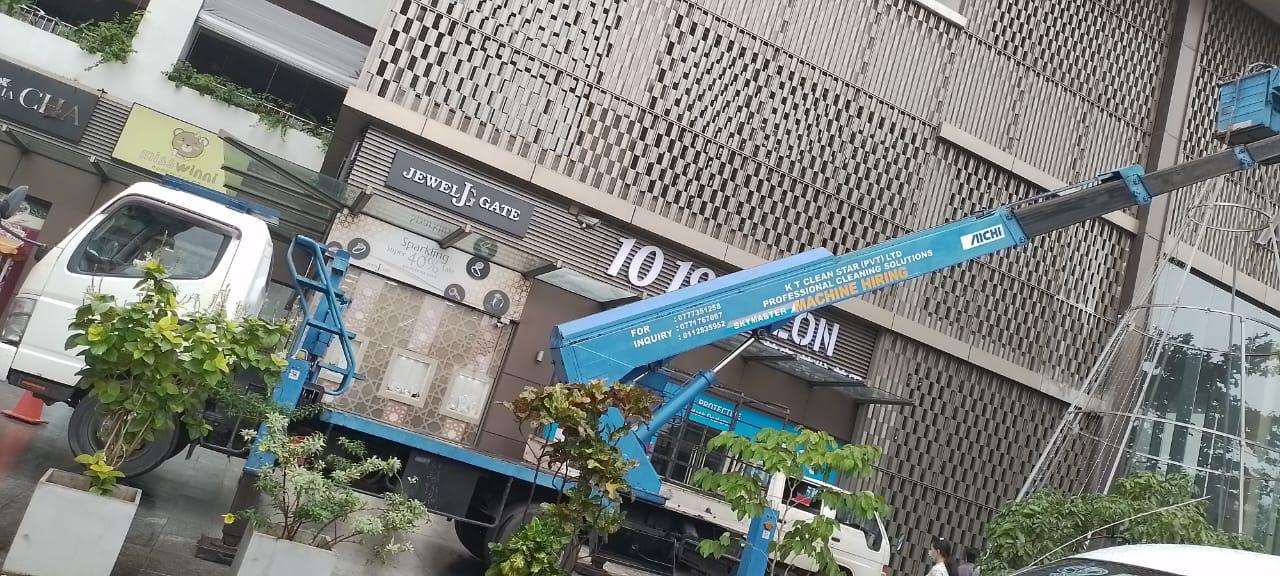 Blue boom lift truck parked in front of a building with plants and glass storefront.