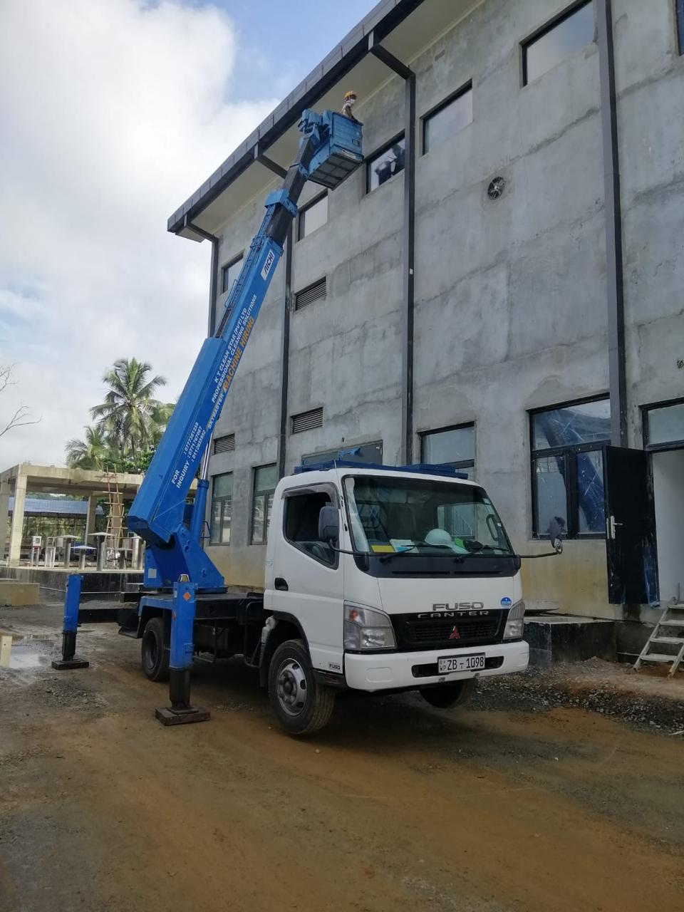 White truck with blue hydraulic lift reaching a worker to the upper wall of a grey building.