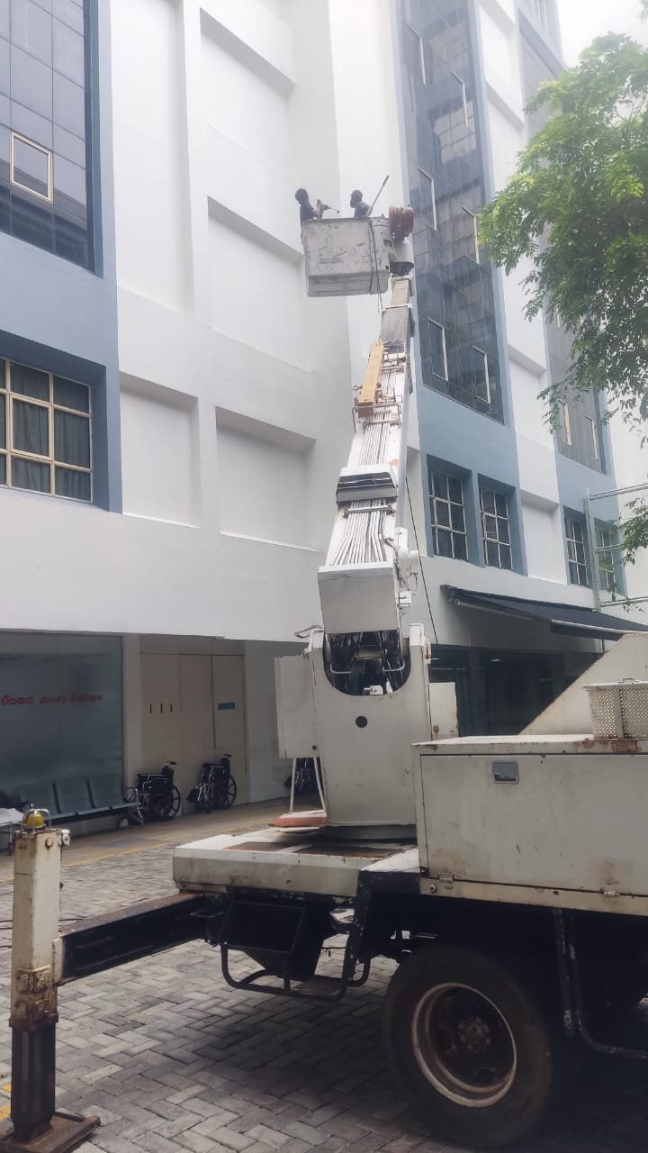 Two workers on elevated platform painting a white building exterior near a tree.