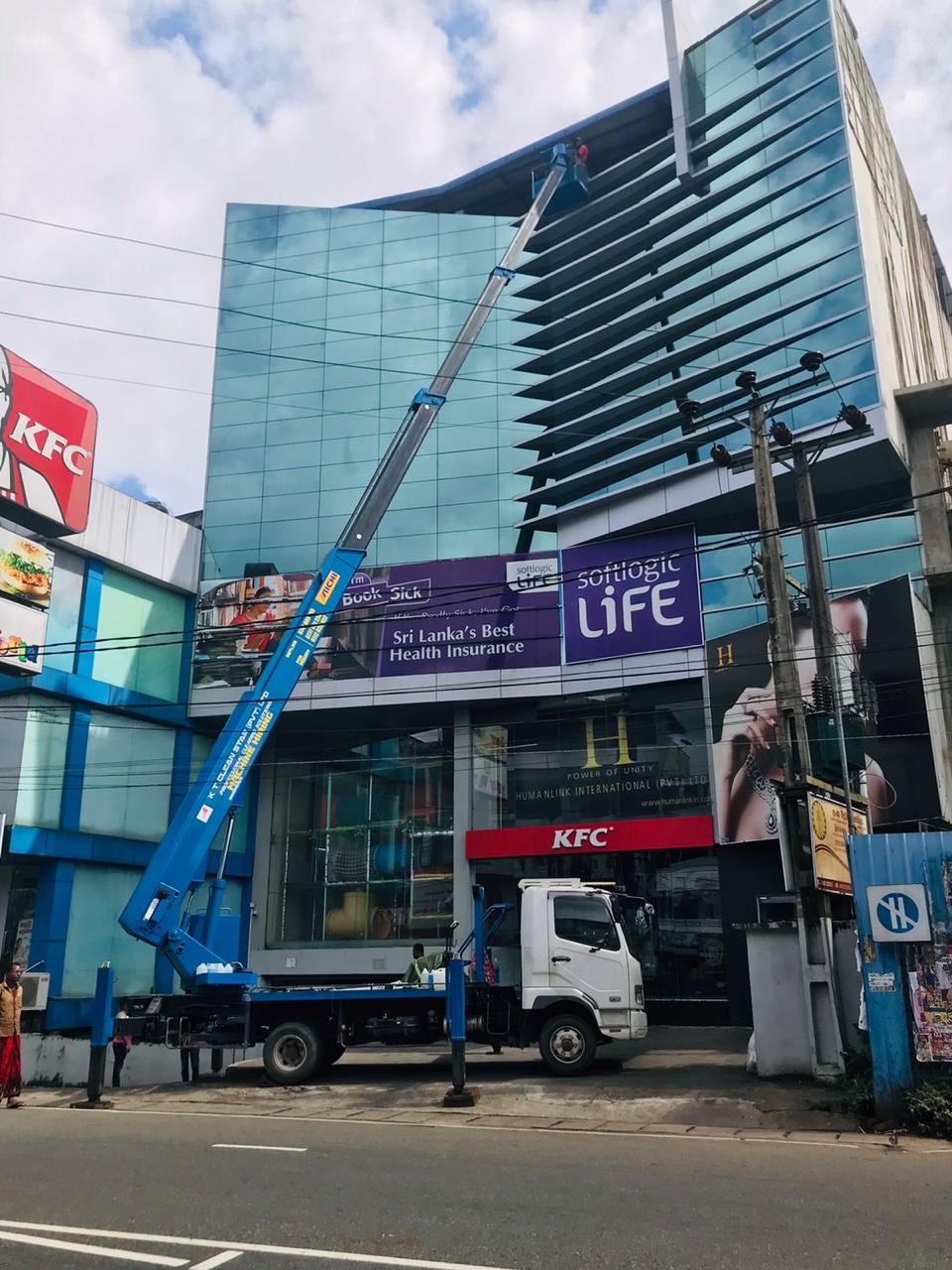 Blue crane truck with extended arm working on glass building front above KFC sign on city street.