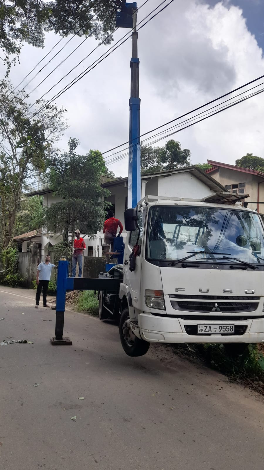 Workers in red shirts use a blue aerial lift on a white truck to reach overhead utility lines by a house.
