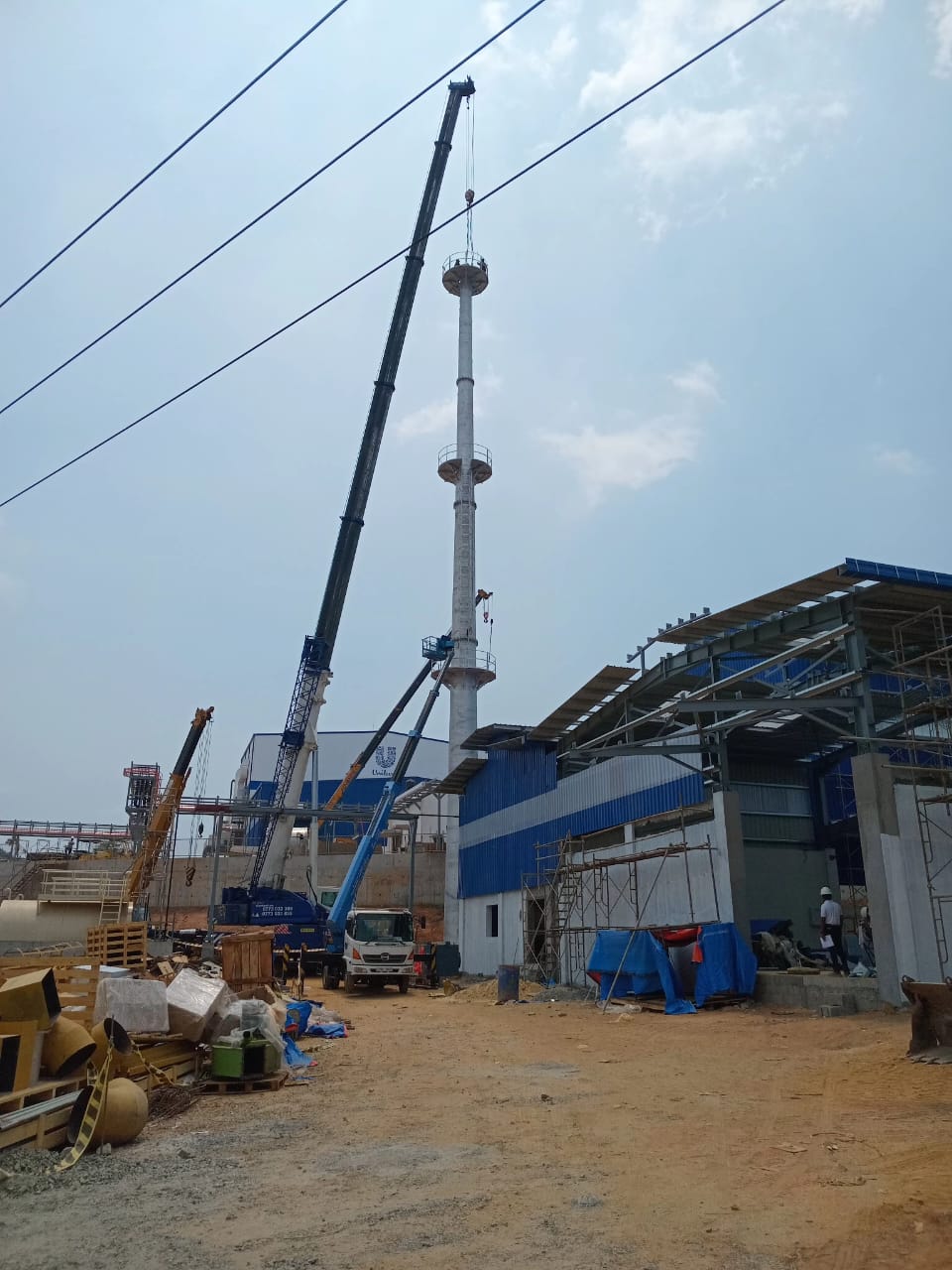 Construction site with cranes assembling a tall industrial chimney near blue and gray buildings.