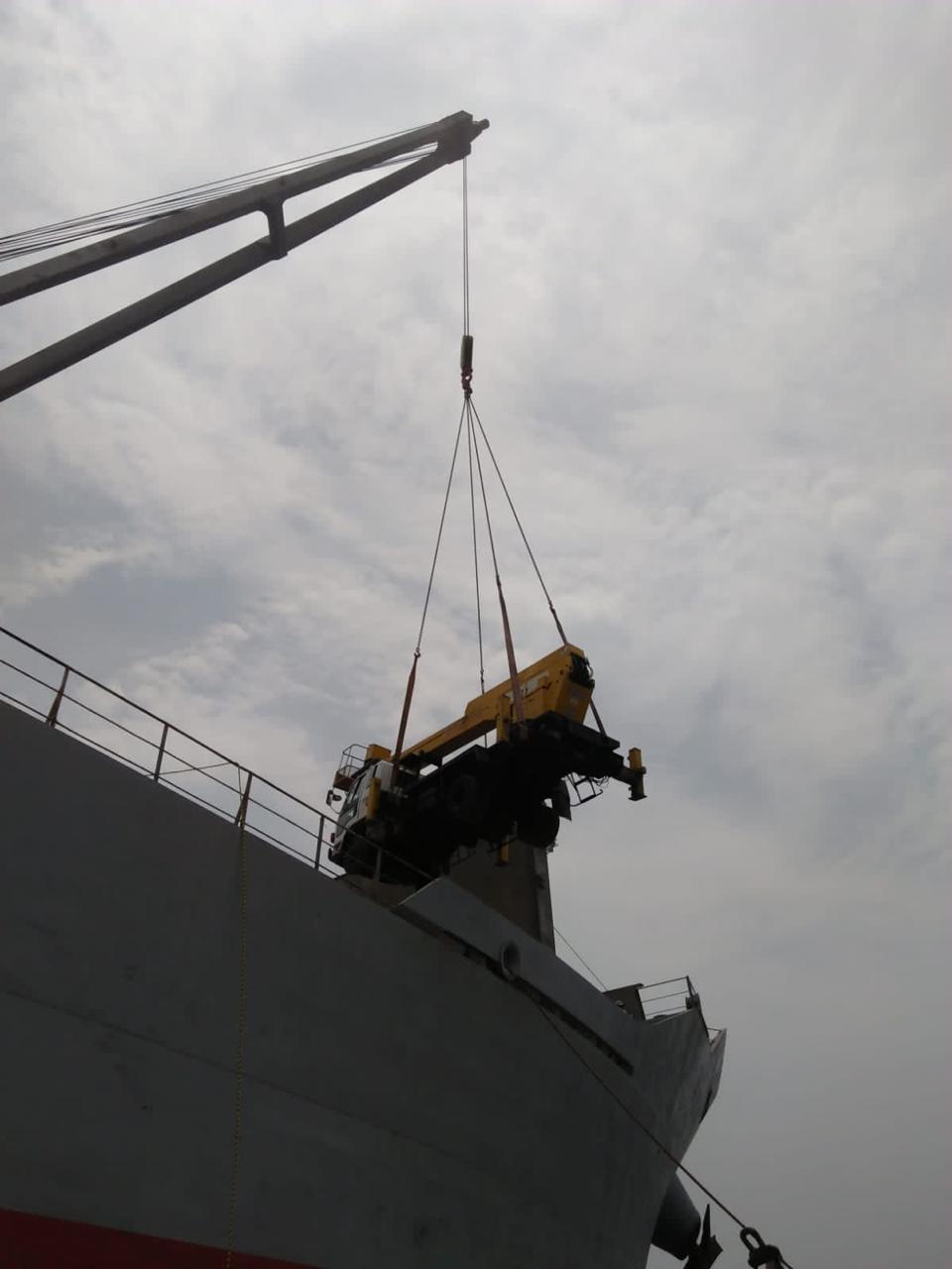 Yellow crane truck being lifted by a ship crane against a cloudy sky.