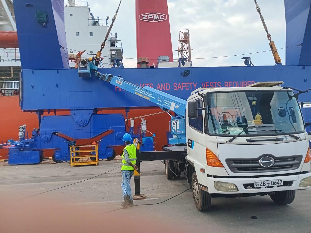 Worker in safety vest and helmet operating a blue hydraulic lift on a white truck near large ship equipment.