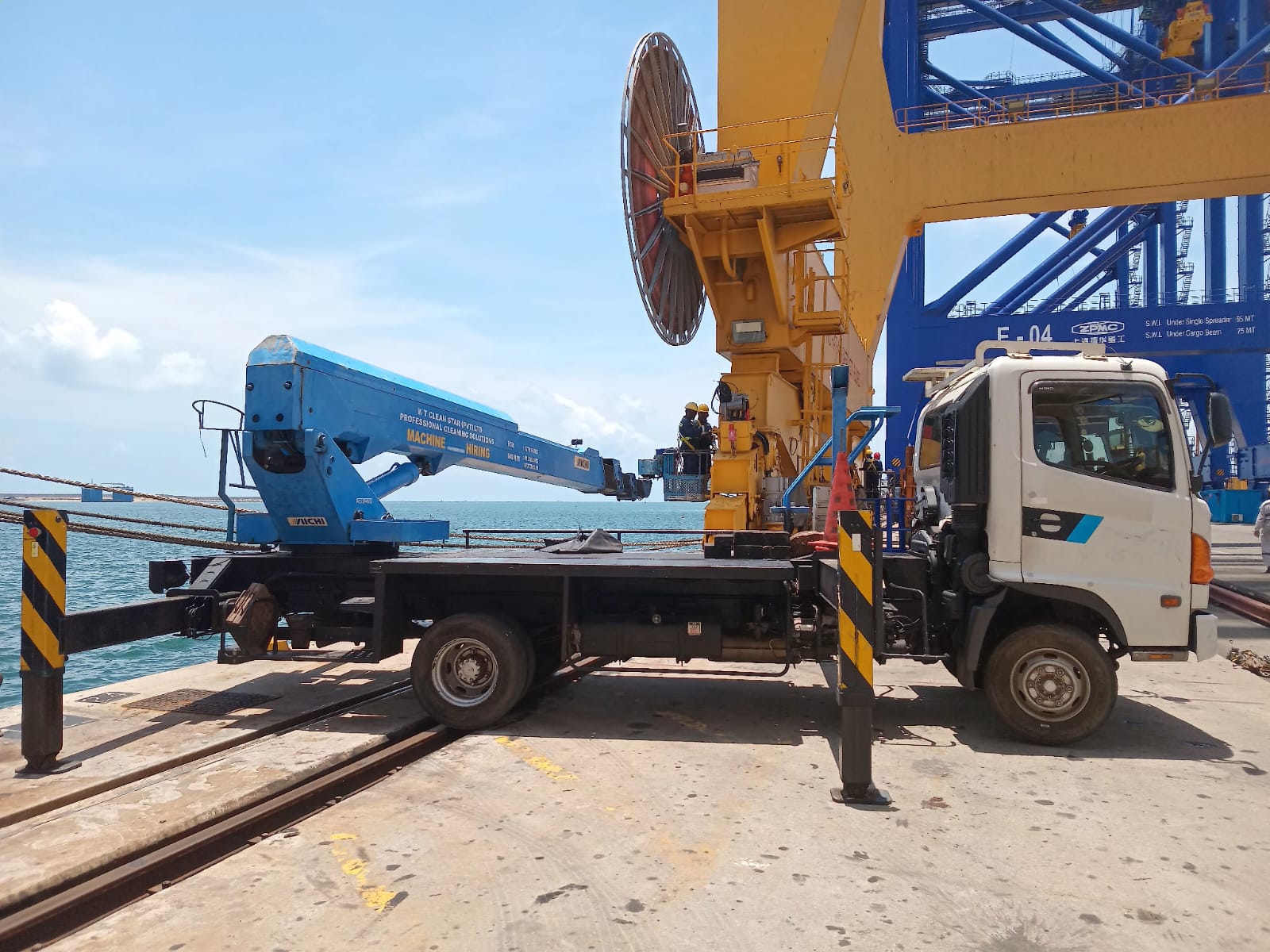 Truck with a blue boom lift parked at a dock with workers in safety gear near a large yellow crane.