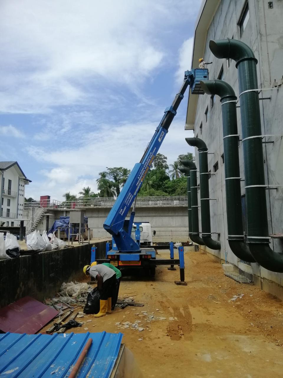 Construction workers with a blue boom lift on site by a building with large black pipes on the wall.