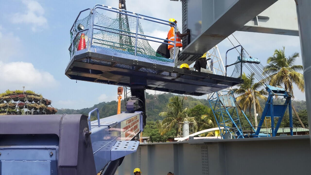 Workers in safety gear on an elevated work platform outdoors near construction equipment and palm trees.