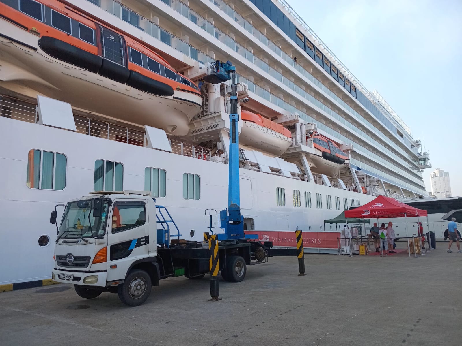 White truck with blue lift parked beside a large Viking cruise ship at a dock with people under a red tent.