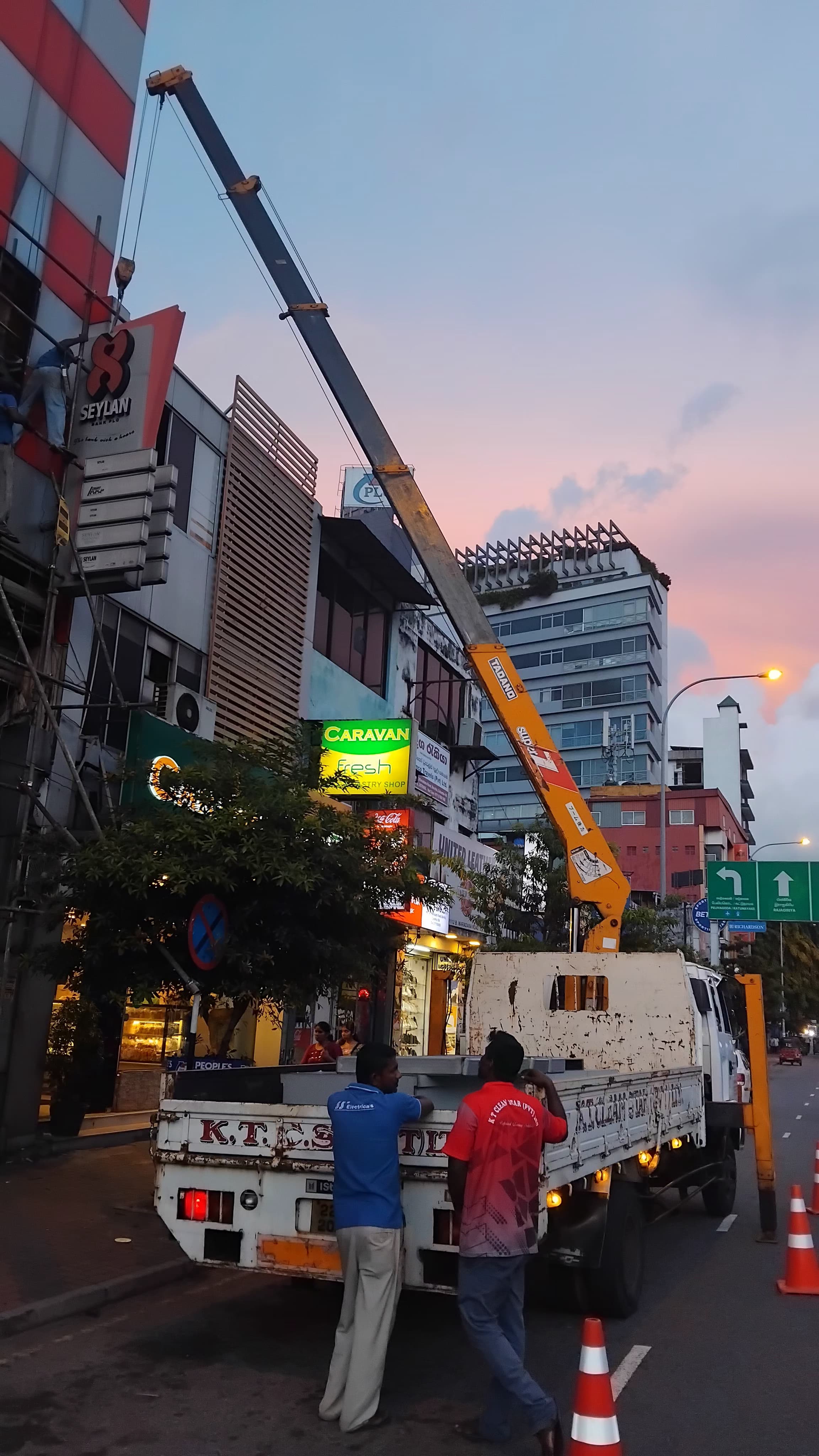 Truck with crane lifting a large Seylan sign on a building during sunset with two men watching nearby.