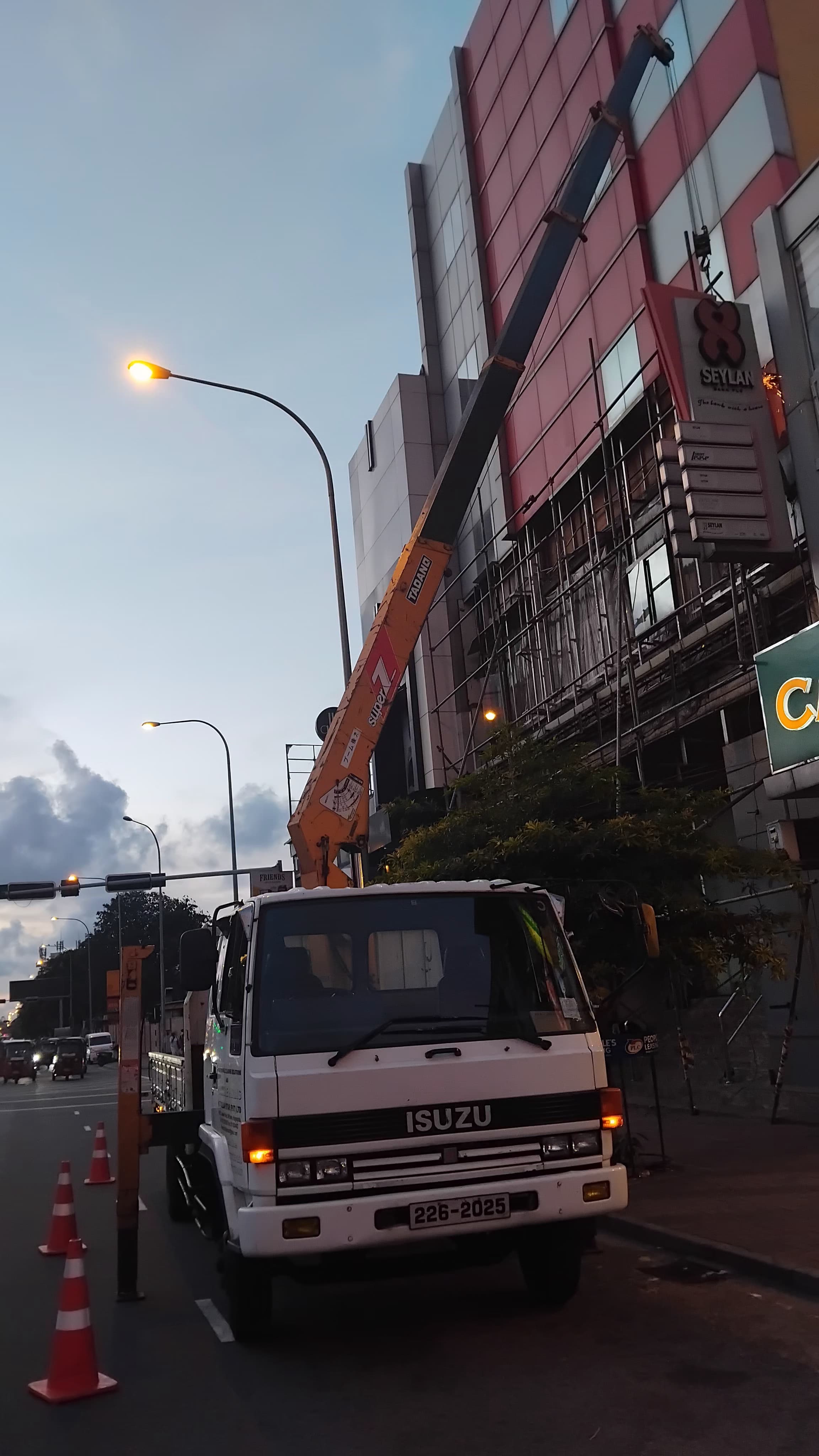 White Isuzu truck with crane extended next to building during dusk, traffic cones on road.