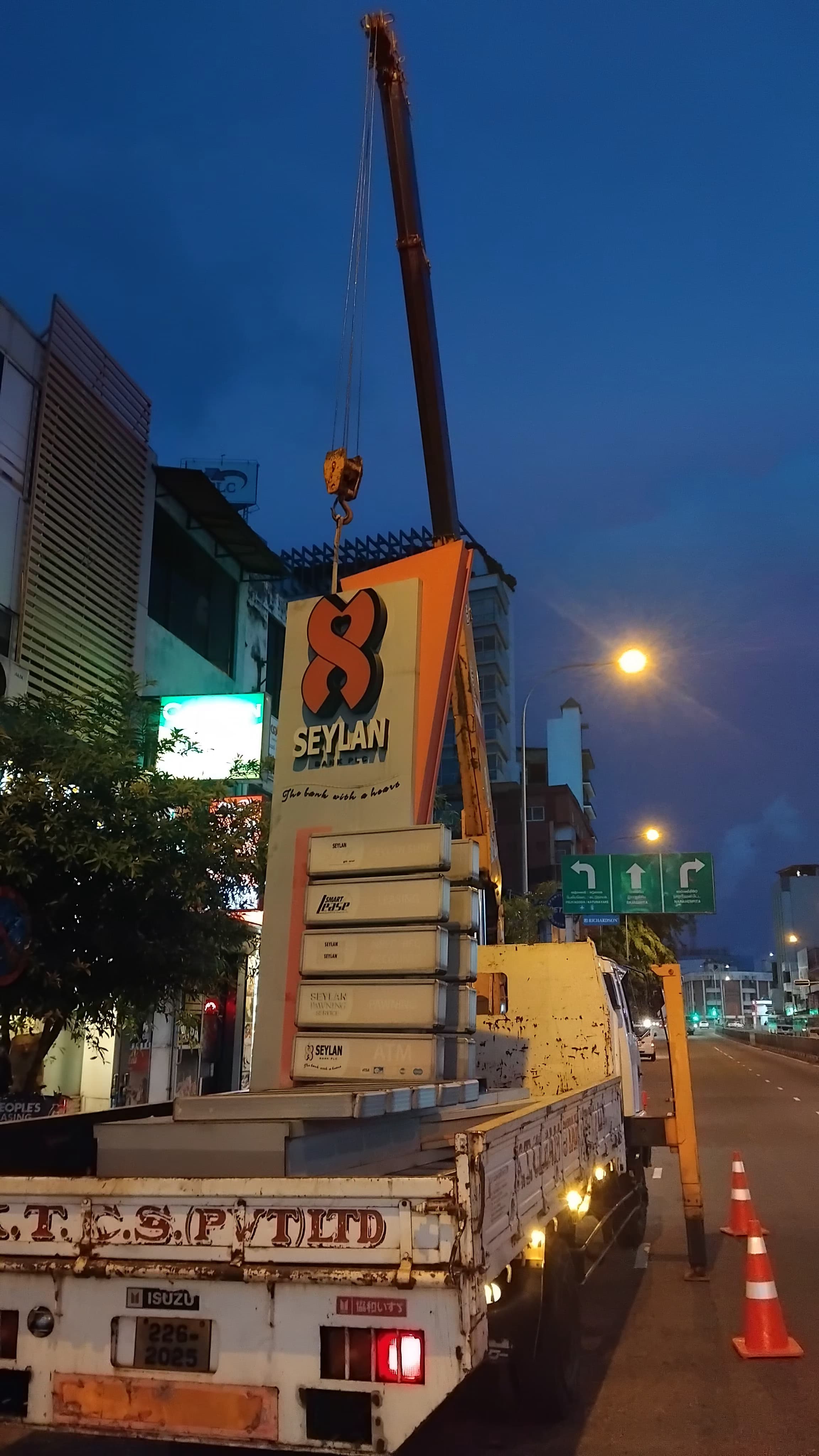 Truck with crane lifting a large illuminated Seylan bank sign on a city street at dusk.
